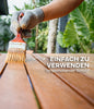 Eine behandschuhte Hand trägt plid Holzöl natürlich & pflegend für Holz innen & außen mit einem Pinsel auf Gartenmöbelbretter auf. Der deutsche Text lautet: EINFACH ZU VERWENDEN, langanhaltender Schutz! Im Hintergrund sind große grüne Blätter zu sehen.