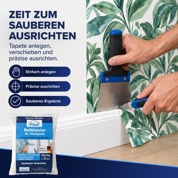 Zwei Hände richten eine Blattmuster-Vliestapete mit Spachtel und Andrückhilfe exakt an der Fußleiste aus, unten links steht der Beutel Plid Rollkleister. Gezeigt wird das Anlegen, Verschieben und präzise Ausrichten der Bahn.