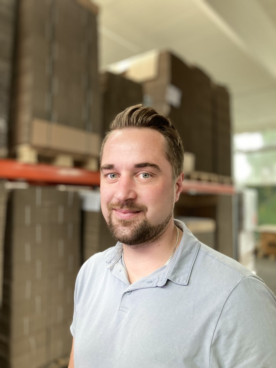Ein Mann mit kurzen braunen Haaren und Bart, der ein hellgraues Polohemd trägt, steht lächelnd in einem Lagerhaus mit Stapeln von Kisten und Regalen im Hintergrund.